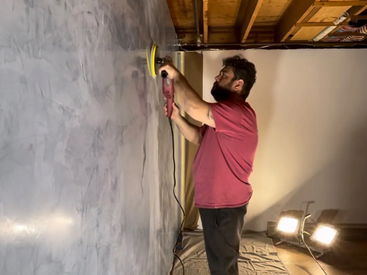 Plasterer burnishing a large grey Venetian plaster wall with a power polisher under work lights